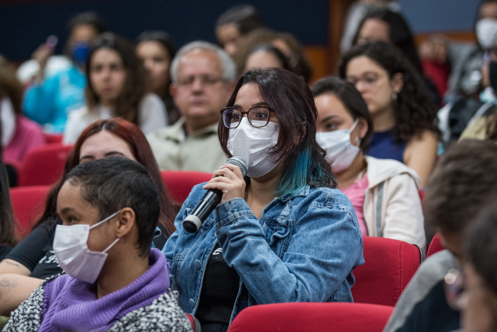 Aluna sentada nas cadeiras no meio do Teatro falando ao microfone.