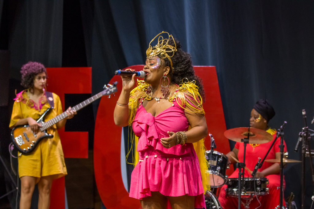 Mulher usando um vestido rosa e adornos dourados canta ao microfone, com o restante da banda e logo da FCA no fundo