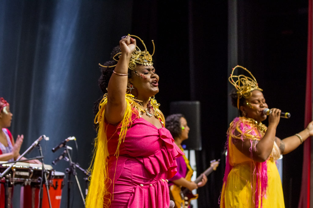 Cantora usando vestido rosa com punho para cima, fazendo sinal de resistência, com banda ao fundo