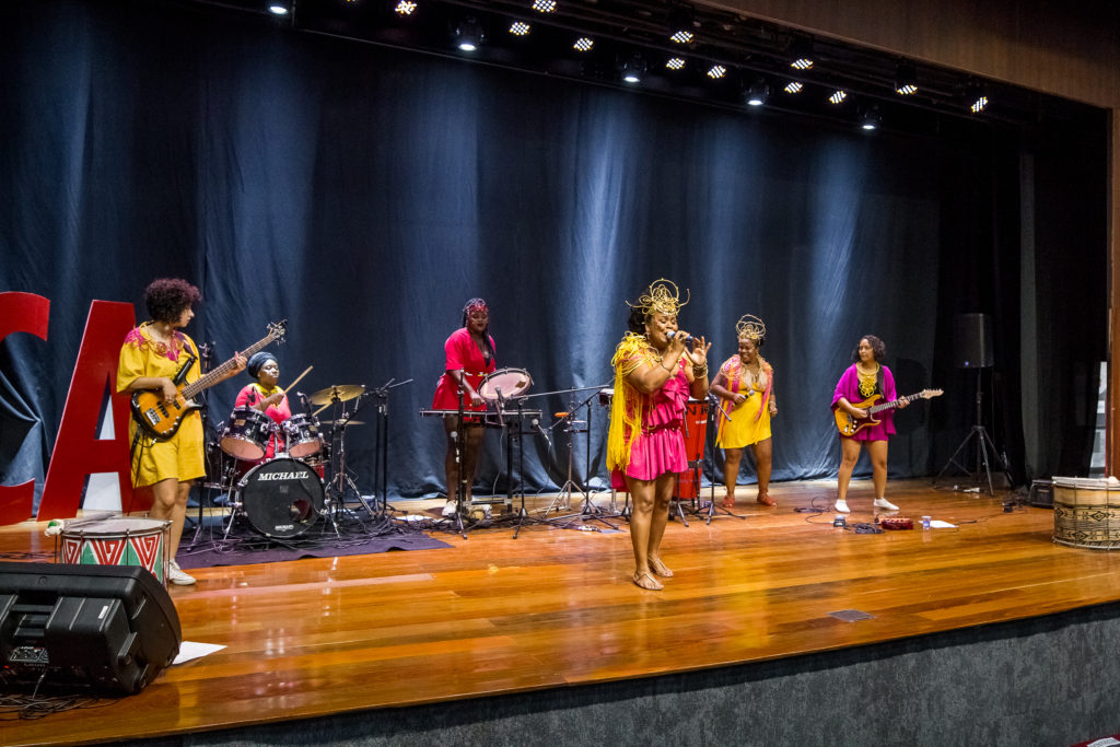 Banda disposta no palco com mulheres usando roupas vermelhas, amarelas e rosas, tocando diversos instrumentos de corda e percussão